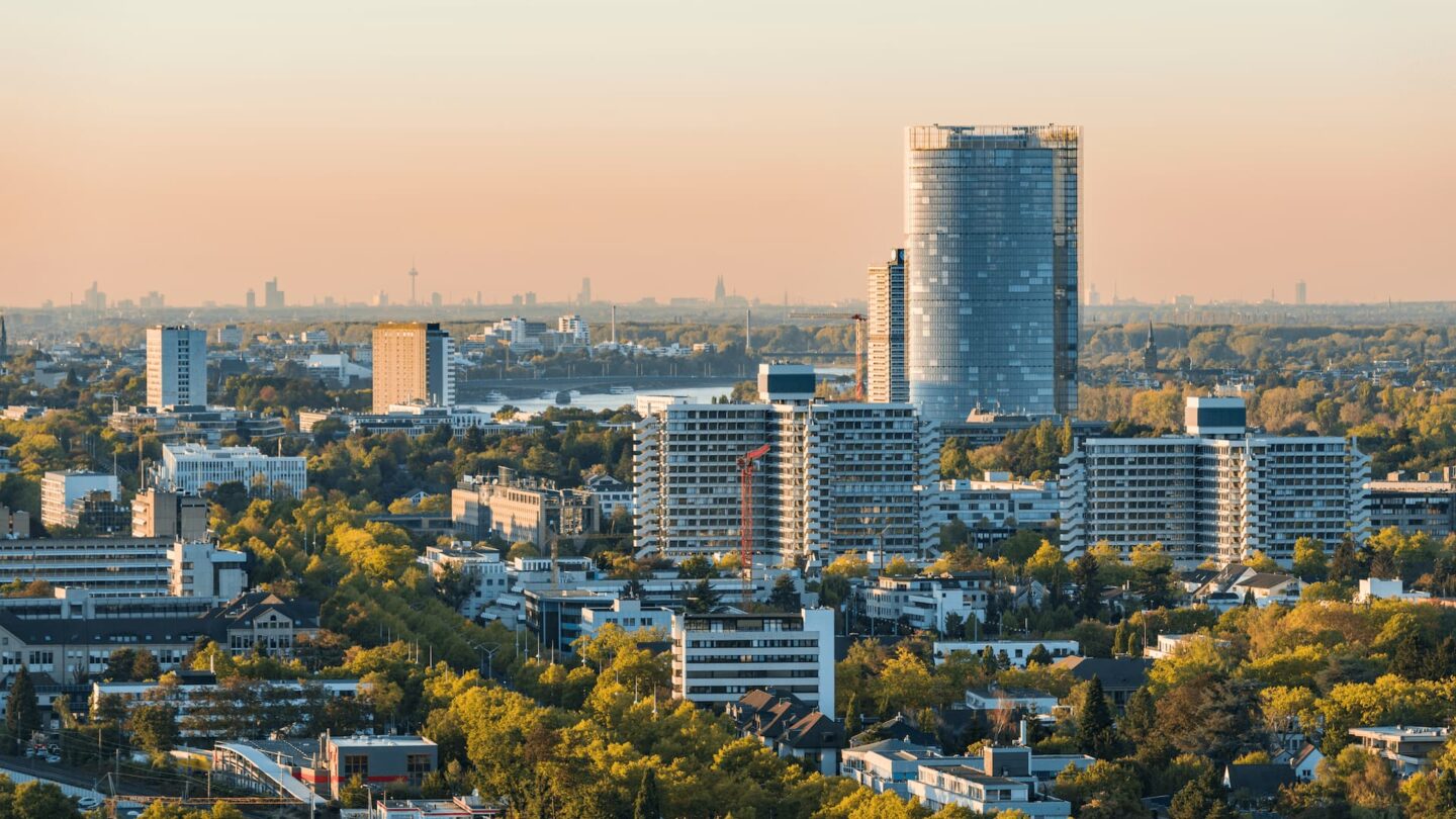 Ein Panoramablick auf Bonn bei Sonnenuntergang, mit einer Mischung aus modernen Hochhäusern und niedrigeren Gebäuden. Im Hintergrund sticht ein markanter Wolkenkratzer hervor, während zwischen den Gebäuden Bäume und Grünflächen verstreut sind. Der Himmel ist klar und spiegelt einen warmen, goldenen Farbton über dieser bezaubernden deutschen Stadtlandschaft.