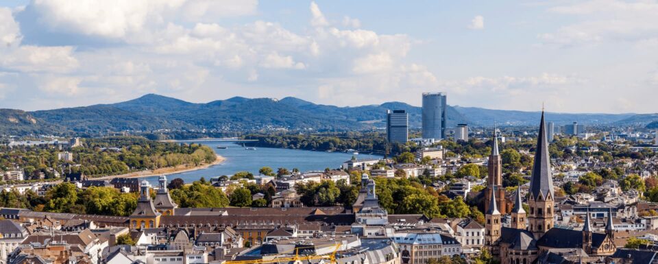 Luftaufnahme von Bonn mit einem durch die Stadt fließenden Fluss, umgeben von Gebäuden, grünen Bäumen und sanften Hügeln im Hintergrund. Moderne Wolkenkratzer und historische Kirchtürme sind sichtbar, mit einer Mischung aus städtischen und natürlichen Landschaften unter einem teilweise bewölkten Himmel.