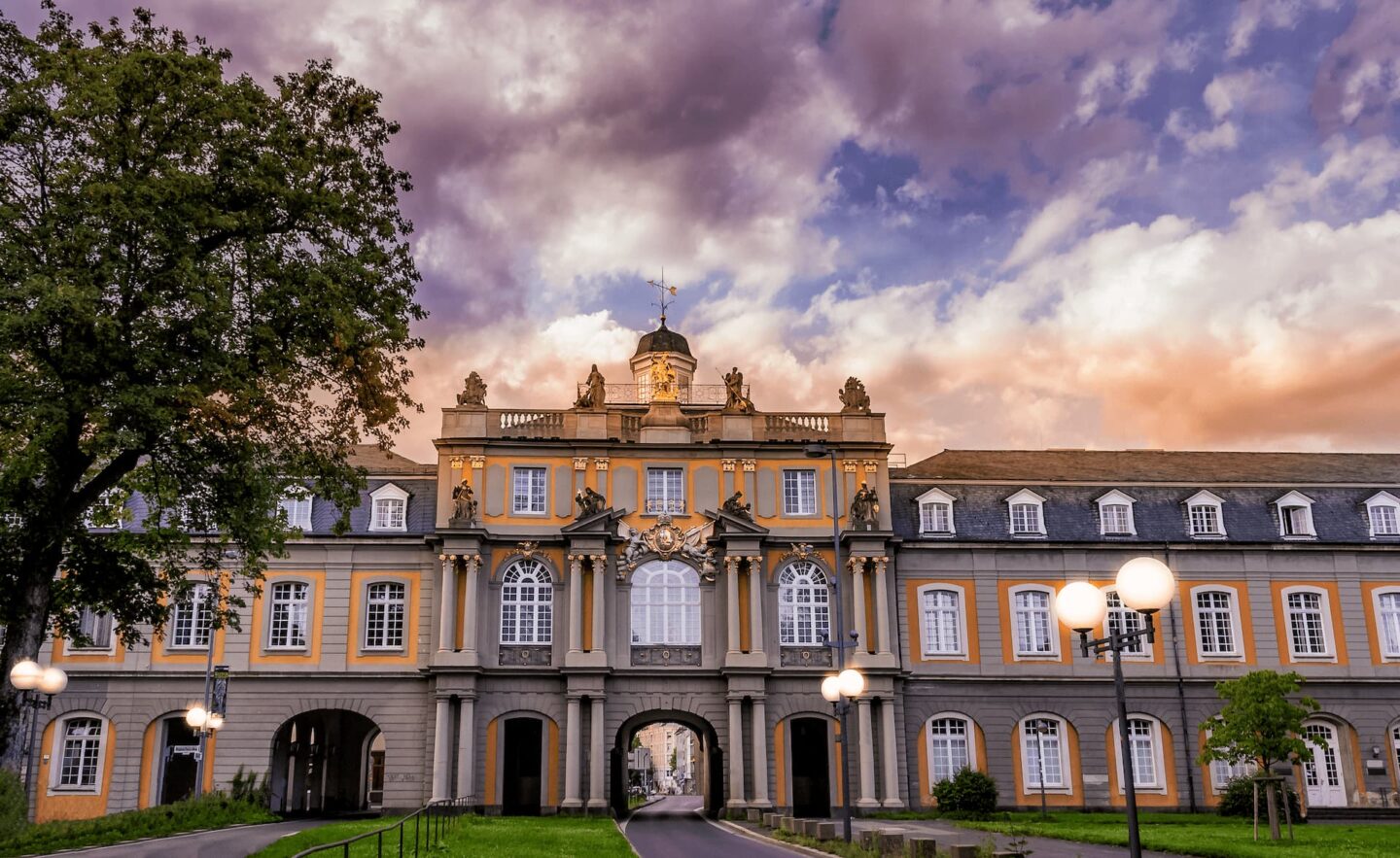 Ein prachtvolles historisches Gebäude mit kunstvoller Architektur, großen Bogenfenstern, dekorativen Statuen und einem zentralen Uhrturm steht vor dem Hintergrund eines dramatischen, farbenfrohen Himmels bei Sonnenuntergang in Bonn. Im Vordergrund sind ein Baum und Laternenpfähle zu sehen.