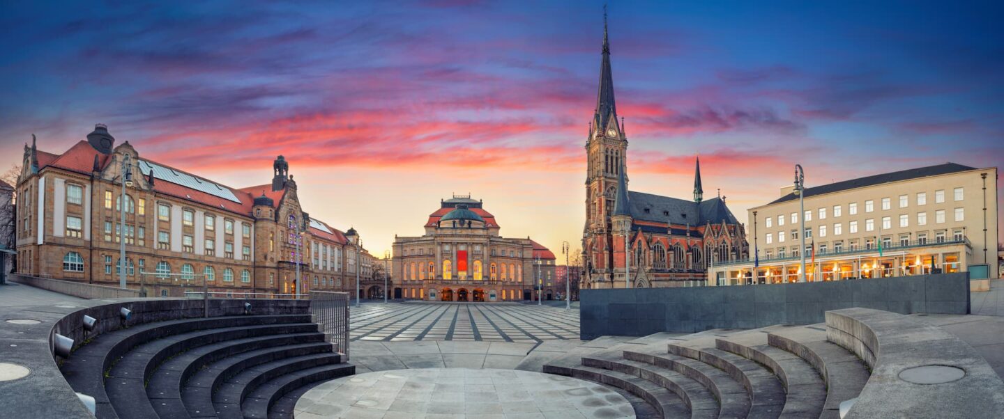 Ein Panoramablick auf einen öffentlichen Platz bei Sonnenuntergang in Chemnitz. Der Platz verfügt im Vordergrund über einen runden, abgestuften Sitzbereich. Um den Platz herum befinden sich historische Gebäude, darunter ein großes Theater in der Mitte und eine hohe Kirche mit einem spitzen Turm auf der rechten Seite.