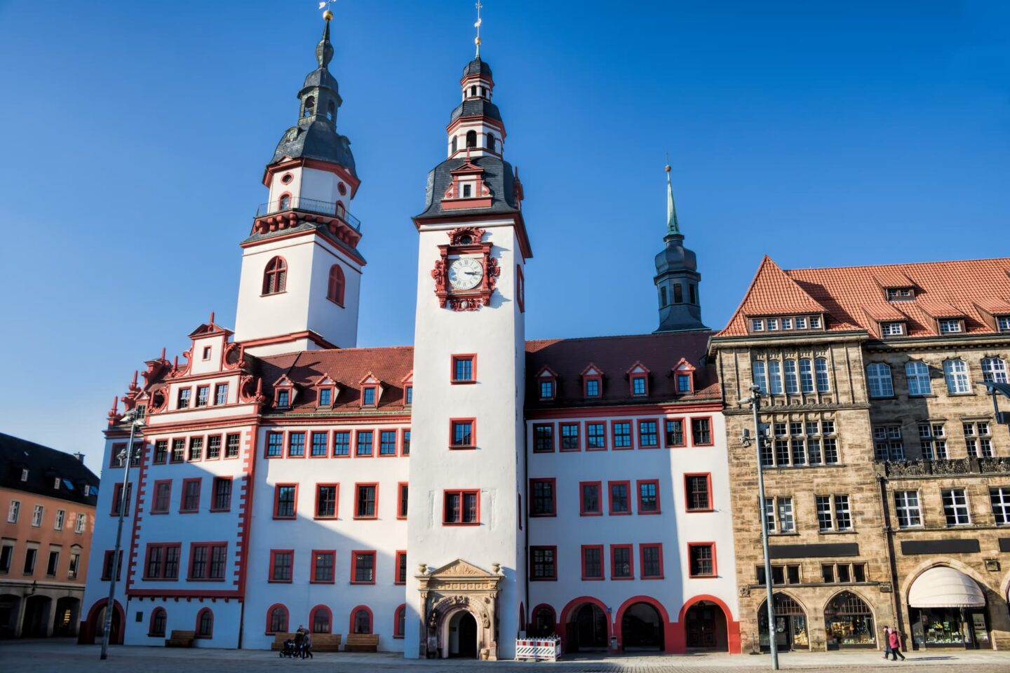 Ein historisches Gebäude in Chemnitz, Sachsen, mit rot-weißen Fassaden, mehreren Türmen mit verzierten Spitzen und einer zentralen Uhr. Die Struktur steht vor einem klaren blauen Himmel. Das Gebäude weist architektonische Details auf und ist von einem Platz umgeben, in dessen Nähe sich einige Menschen aufhalten.