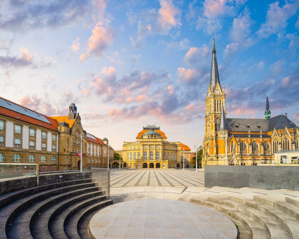 Ein malerischer europäischer Stadtplatz bei Sonnenuntergang in Chemnitz, mit einer großen Kirche mit einem hohen Turm auf der rechten Seite, historischen Gebäuden mit orangefarbenen Dächern und kunstvollen Fassaden auf der linken Seite und Treppen im Vordergrund. Der Himmel ist mit flauschigen rosa Wolken gefüllt.