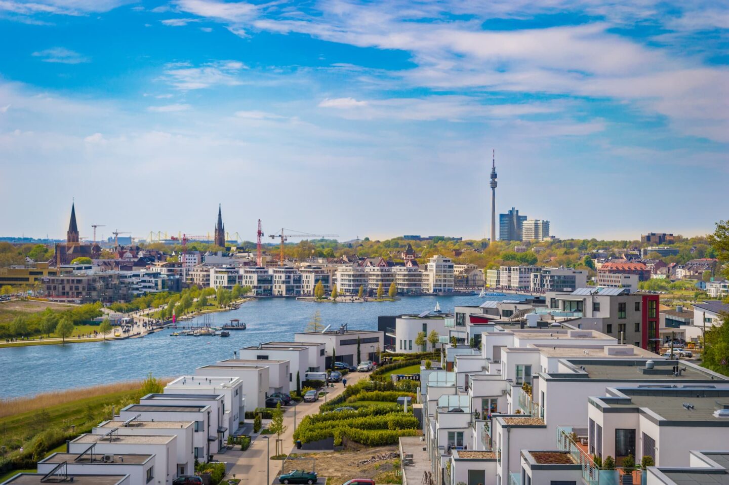 Eine Stadtlandschaft mit modernen weißen Wohnhäusern mit Flachdächern im Vordergrund, einem Fluss, der durch die Mitte fließt, und einer Skyline mit Kirchtürmen und einem hohen Turm im Hintergrund vor einem teilweise bewölkten blauen Himmel erinnert an den Charme Dortmunds.