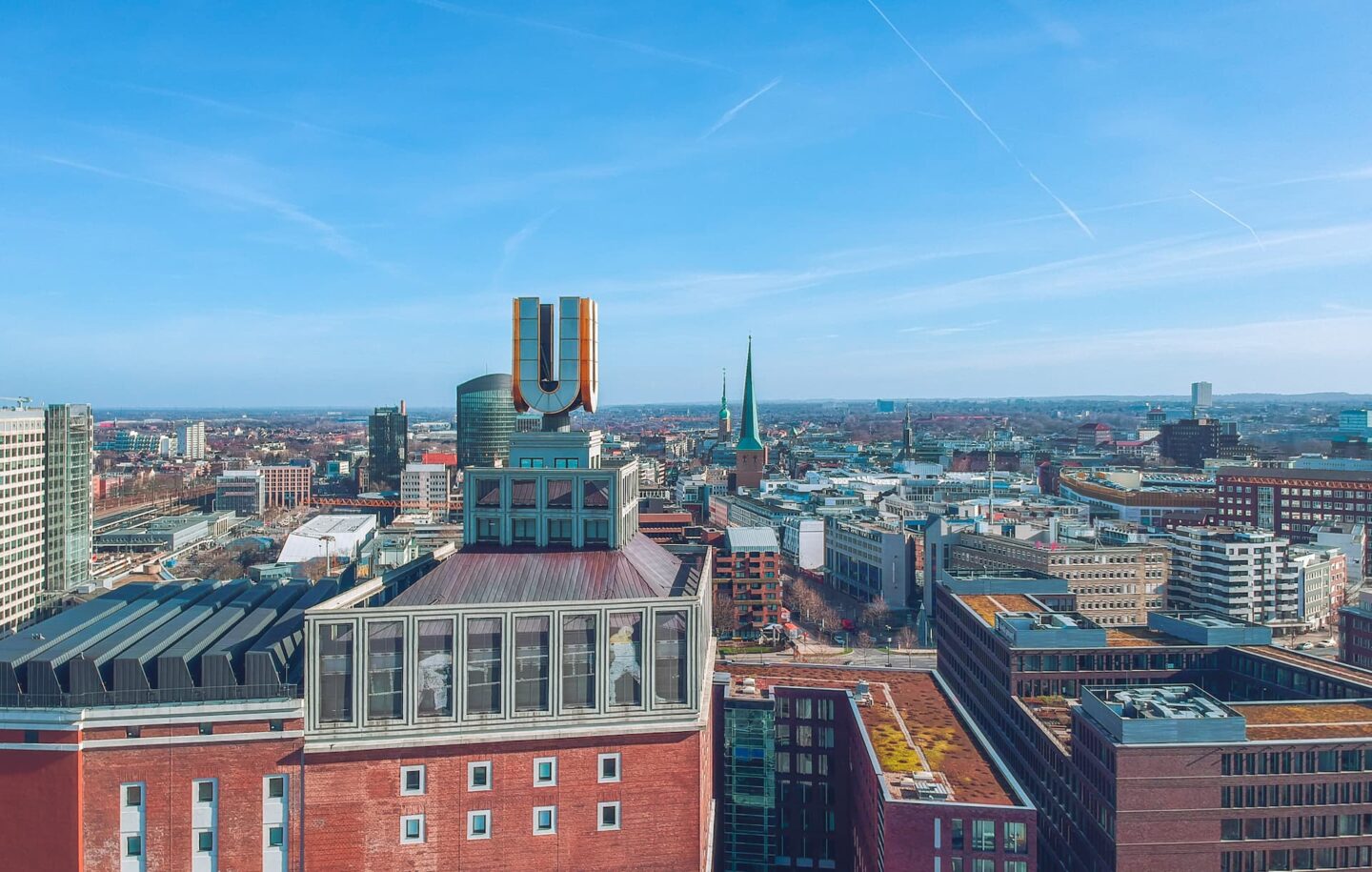 Ein Panoramablick auf die Stadtlandschaft Dortmunds an einem klaren Tag. Im Vordergrund ist ein großes, markantes rotes Backsteingebäude mit einer markanten „U“-Skulptur zu sehen. Die Stadt ist dicht mit verschiedenen Gebäuden bebaut und in der Mitte ist ein Kirchturm zu sehen.