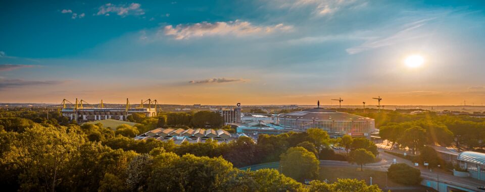 Ein Panoramablick auf Dortmund bei Sonnenuntergang zeigt eine Mischung aus moderner und traditioneller Architektur. Die Skyline umfasst ein Stadion mit Kränen im Hintergrund. Im Vordergrund ist eine üppige Grünfläche mit Bäumen zu sehen, die der städtischen Umgebung dieser deutschen Stadt natürliche Schönheit verleiht – ein Genuss für Reisende.