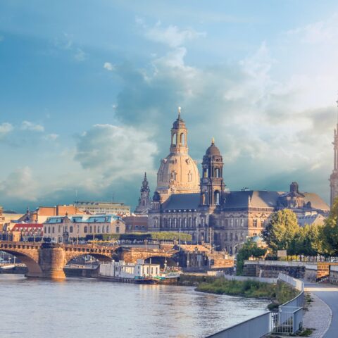 Eine malerische Aussicht auf Dresden, Deutschland. Das Bild zeigt historische Architektur entlang der Elbe, darunter die Frauenkirche und den Dresdner Dom. Der Himmel ist teilweise bewölkt und die Sonne beleuchtet die Gebäude. Bäume säumen den Uferweg – ein perfektes Ziel für Reiseliebhaber, die reiche Geschichte und Schönheit suchen.