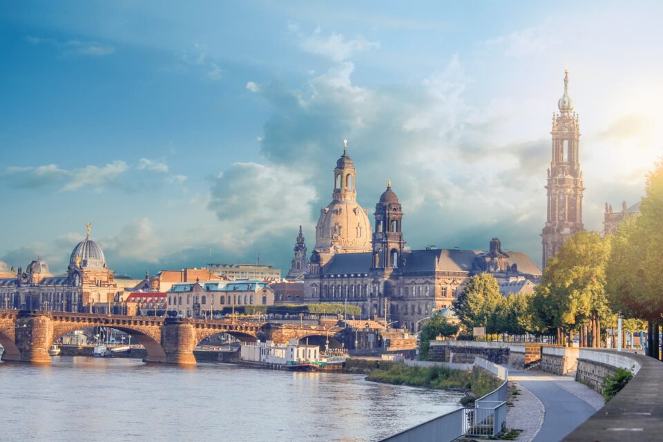 Eine malerische Aussicht auf Dresden, Deutschland. Das Bild zeigt historische Architektur entlang der Elbe, darunter die Frauenkirche und den Dresdner Dom. Der Himmel ist teilweise bewölkt und die Sonne beleuchtet die Gebäude. Bäume säumen den Uferweg – ein perfektes Ziel für Reiseliebhaber, die reiche Geschichte und Schönheit suchen.