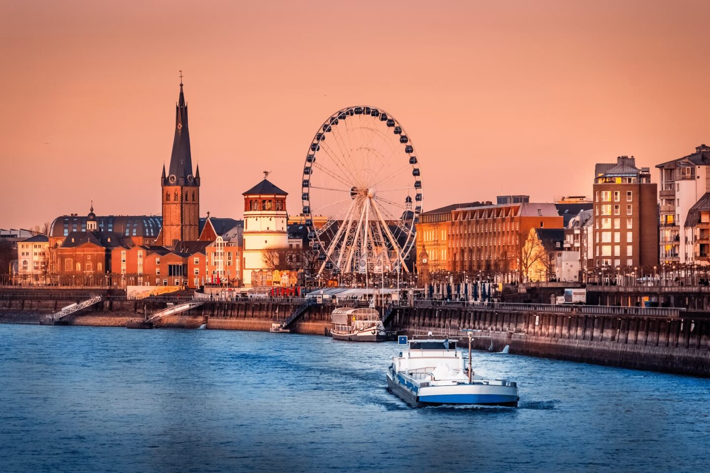 Eine ruhige Stadtlandschaft am Wasser bei Sonnenuntergang. Im Vordergrund ist eine Wasserfläche zu sehen, auf die sich ein Boot nähert. Im Hintergrund sind Düsseldorfs Riesenrad, der Kirchturm und verschiedene Gebäude in warmes Abendlicht getaucht.