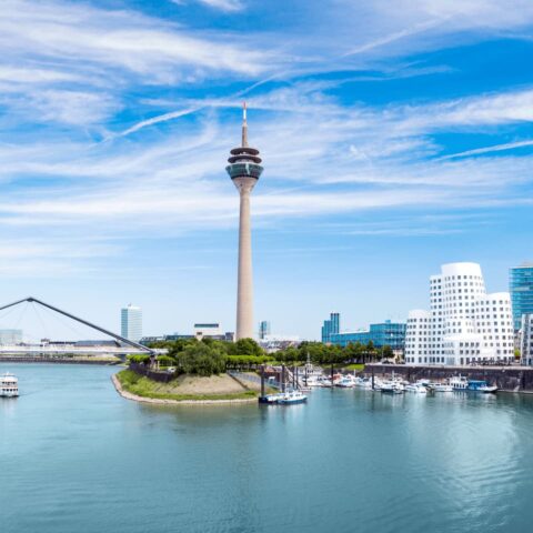 Ein malerischer Blick auf die Skyline von Düsseldorf mit dem Rheinturm in der Mitte. Moderne Gebäude, darunter die ikonischen Gehry-Bauten, säumen die Uferpromenade. Am Fluss liegen Boote vor Anker, über uns ein klarer blauer Himmel.