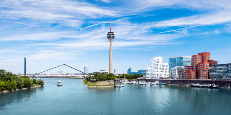 Ein malerischer Blick auf die Skyline von Düsseldorf mit dem Rheinturm in der Mitte. Moderne Gebäude, darunter die ikonischen Gehry-Bauten, säumen die Uferpromenade. Am Fluss liegen Boote vor Anker, über uns ein klarer blauer Himmel.