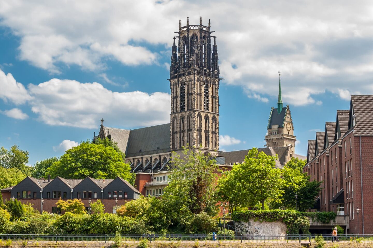 Eine malerische Aussicht auf Duisburg mit einem hohen Dom im gotischen Stil und einem markanten Turm, umgeben von verschiedenen Gebäuden, üppigen grünen Bäumen und einem teilweise bewölkten blauen Himmel.