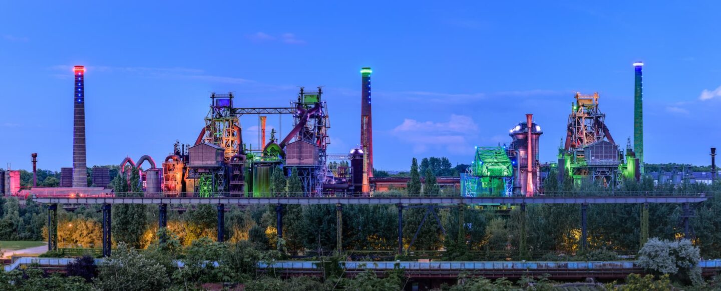 Ein Panoramablick auf einen großen Industriekomplex in Duisburg bei Dämmerung zeigt mehrere alte Fabrikgebäude und Türme, die in leuchtenden Farben wie Grün, Blau und Lila beleuchtet sind. Im Vordergrund sind grüne Bäume und Vegetation zu sehen, während der Himmel einen Blauverlauf aufweist.