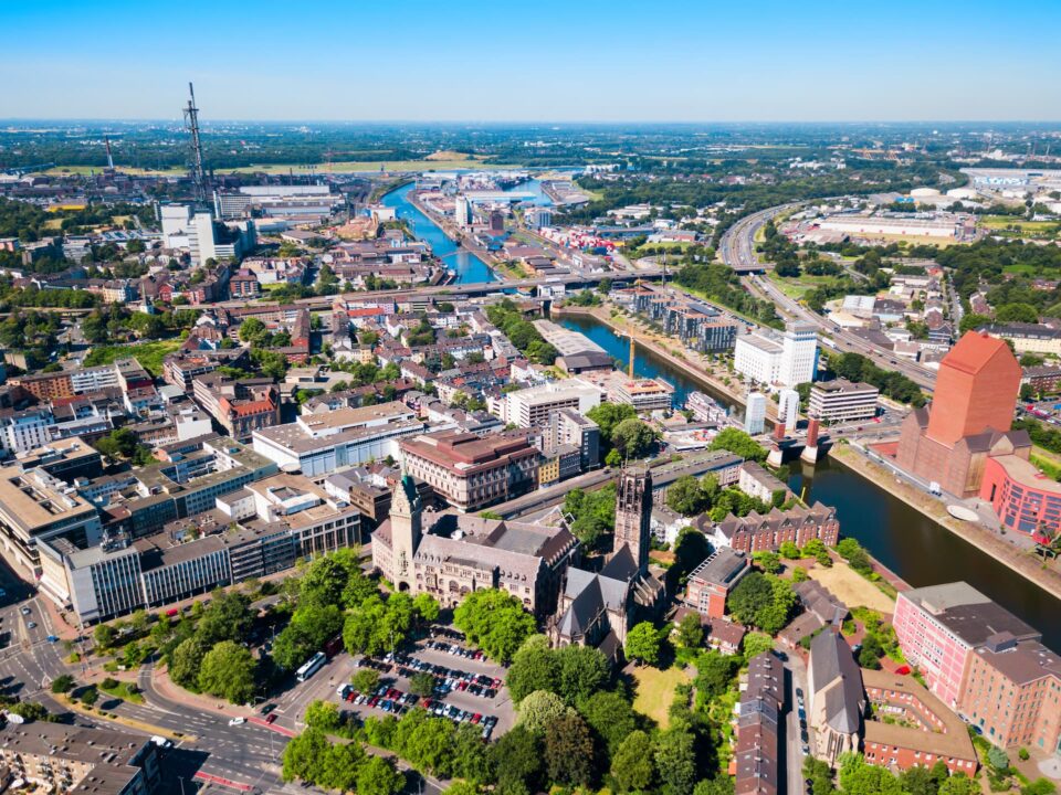 Luftaufnahme von Duisburg, die eine Mischung aus modernen Gebäuden und historischer Architektur zeigt. Im Vordergrund ist eine markante Kirche von Grün umgeben, durch die Mitte verlaufen ein Fluss und mehrere Brücken. Straßen und Infrastruktur sind im gesamten Gebiet sichtbar.