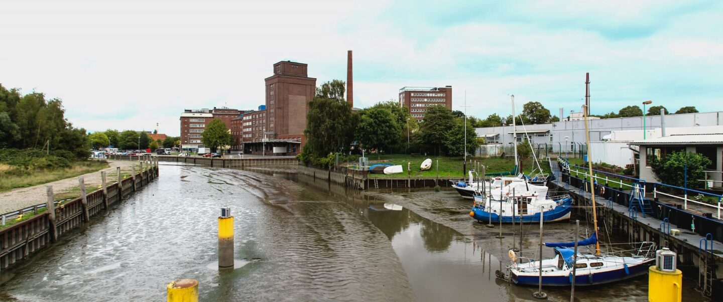 Ein kleiner Hafen in Elmshorn mit Booten, die bei Ebbe angedockt sind und Watt und seichtes Wasser freigeben. Das Ufer ist gesäumt von grünen Bäumen, Gebäuden und Industrieanlagen. Ein bedeckter Himmel trägt zur ruhigen Atmosphäre bei. Im schlammigen Wasser sind gelbe Anlegestellen zu sehen.