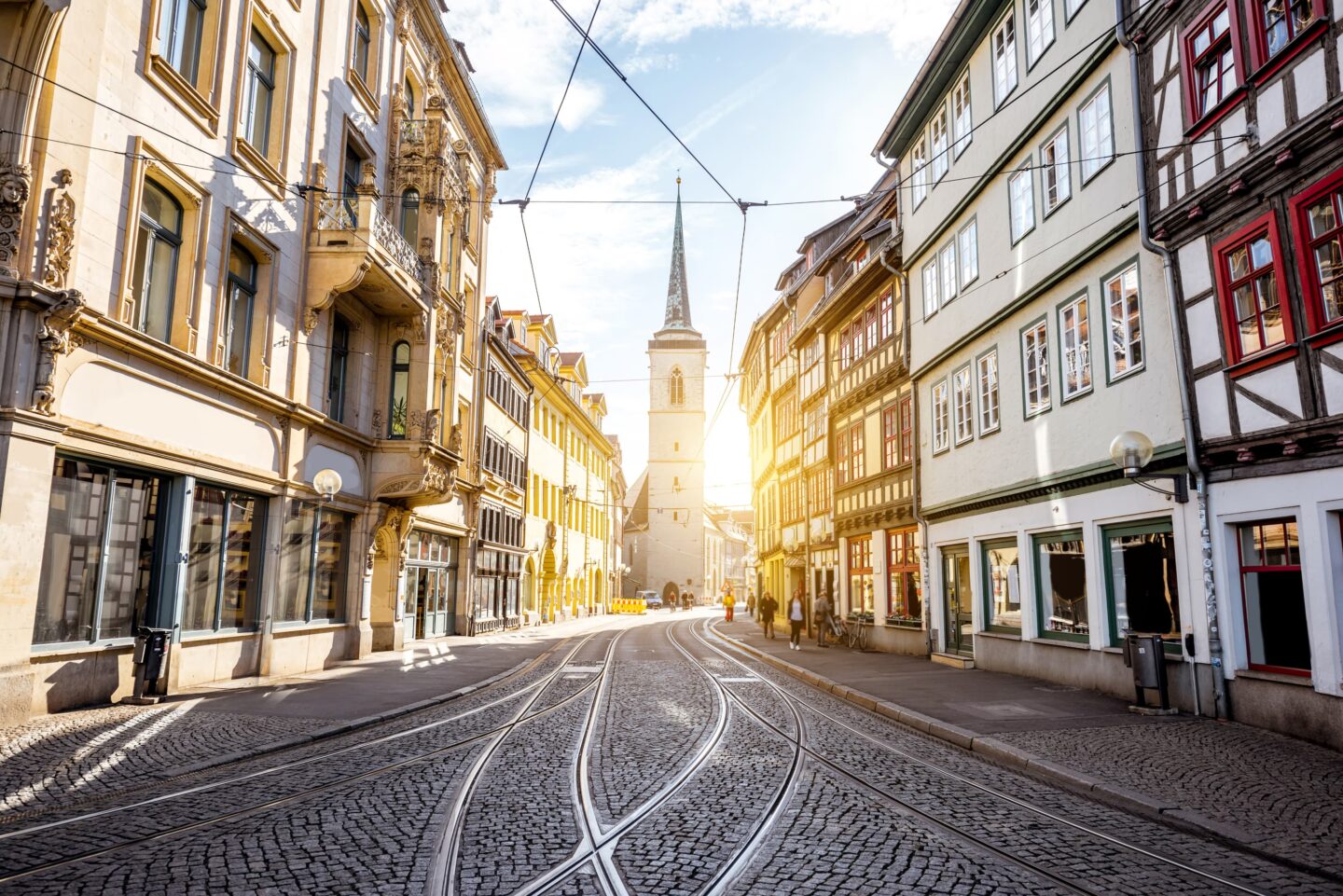 Eine malerische europäische Straßenszene in Erfurt mit farbenfrohen historischen Gebäuden entlang einer Kopfsteinpflasterstraße, die auf einen entfernten Kirchturm zuläuft. Das warme Sonnenlicht schafft eine charmante und einladende Atmosphäre und hebt die Architektur und die Straßenbahnschienen im Vordergrund hervor.