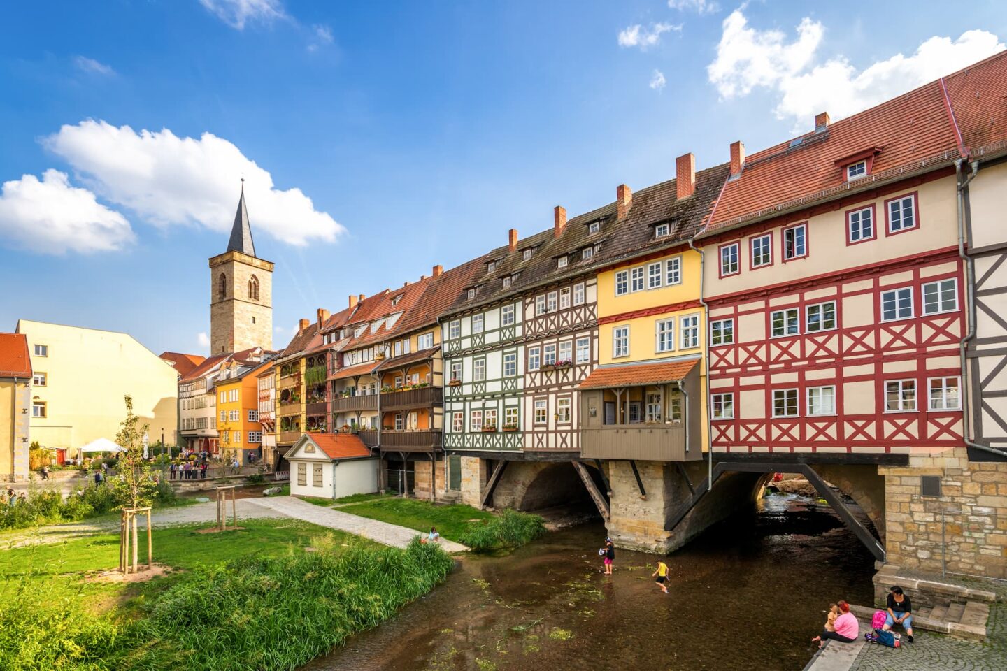 Eine malerische Szene der Krämerbrücke in Erfurt. Die historischen Fachwerkhäuser erstrecken sich über beide Seiten eines Flusses und haben leuchtende Fassaden in Rot, Gelb und Beige. Unten sieht man Menschen, die das seichte Wasser und das sonnige Wetter genießen.