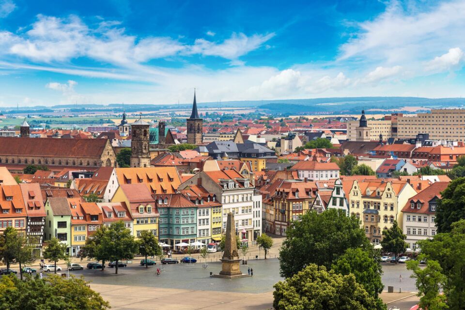 Ein Panoramablick auf die europäische Stadt Erfurt zeigt farbenfrohe historische Gebäude, einen markanten Kirchturm und einen zentralen Platz mit einem kleinen Denkmal. Der Himmel ist strahlend blau mit vereinzelten Wolken. Im Vordergrund sind Bäume zu sehen.