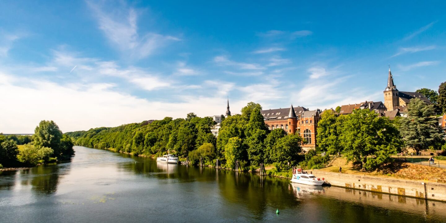 Eine ruhige Flusslandschaft in Essen mit üppigen grünen Bäumen entlang der Ufer. Mehrere Boote liegen am Fluss angedockt und im Hintergrund sind unter einem strahlend blauen Himmel mit vereinzelten Wolken charmante Backsteingebäude zu sehen, darunter eine Kirche mit einem hohen Turm, die zu Reisen und Tourismus einlädt.