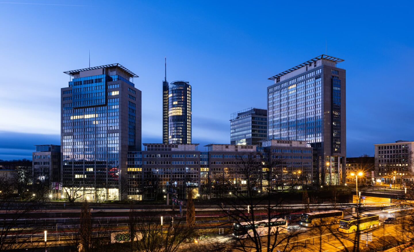 Eine Stadtansicht in der Abenddämmerung in Essen zeigt mehrere moderne Bürogebäude mit Glasfassaden, die vor einem Abendhimmel beleuchtet sind. Die Szene umfasst belebte Straßen mit Lichtspuren von Fahrzeugen und eine Straßenbahnhaltestelle im Vordergrund.