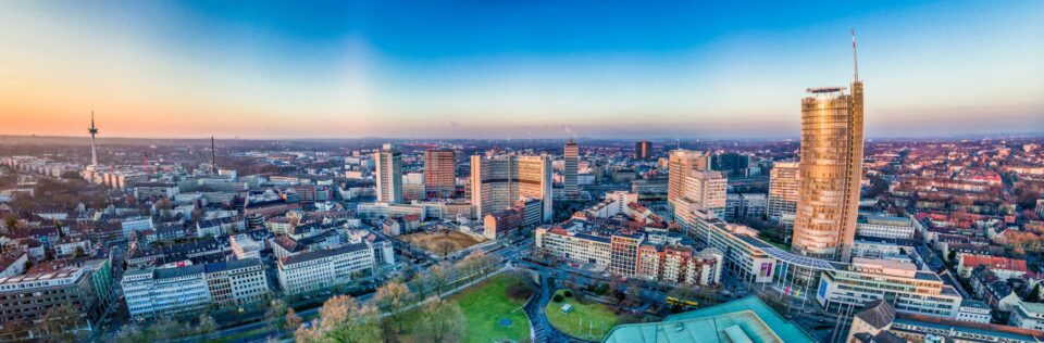 Ein Panoramablick auf die Skyline von Essen bei Sonnenuntergang. Hohe Gebäude, darunter markante Turmstrukturen, dominieren die Szene. Auf den Straßen darunter herrscht reger Verkehr und in der Ferne erstrecken sich Wohngebiete unter einem leuchtend blauen und orangefarbenen Himmel.