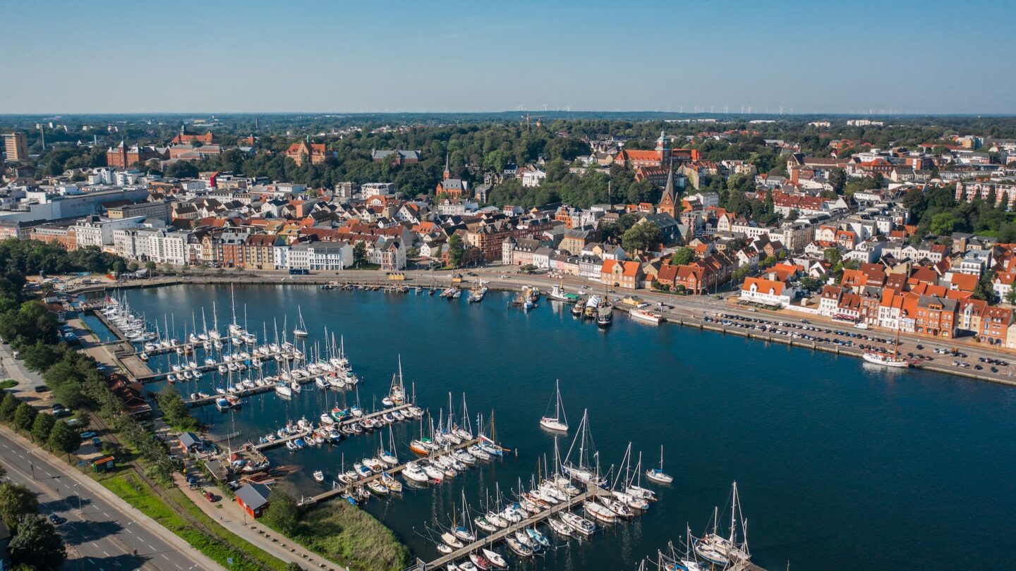 Luftaufnahme von Flensburg, einer Küstenstadt mit einem geschäftigen Yachthafen, an dessen Piers zahlreiche Boote ankern. Die Uferpromenade ist von farbenfrohen Gebäuden gesäumt, und eine Mischung aus Wohn- und Geschäftshäusern erstreckt sich in Richtung einer grünen, bewaldeten Gegend in der Ferne.