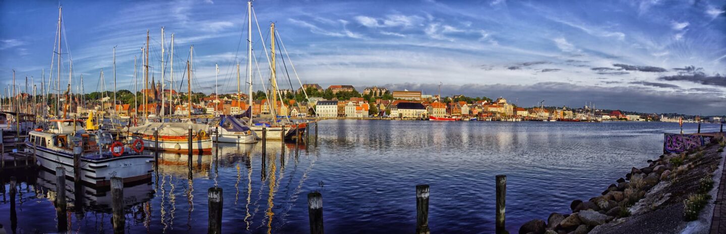 Ein Panoramablick auf den Flensburger Hafen mit angedockten Segelbooten und Motorbooten unter einem teilweise bewölkten Himmel. Am gegenüberliegenden Ufer befindet sich eine dichte Ansammlung farbenfroher Gebäude, im Hintergrund etwas Grün auf einem Hügel. Das ruhige Wasser spiegelt die Boote und Gebäude wider.