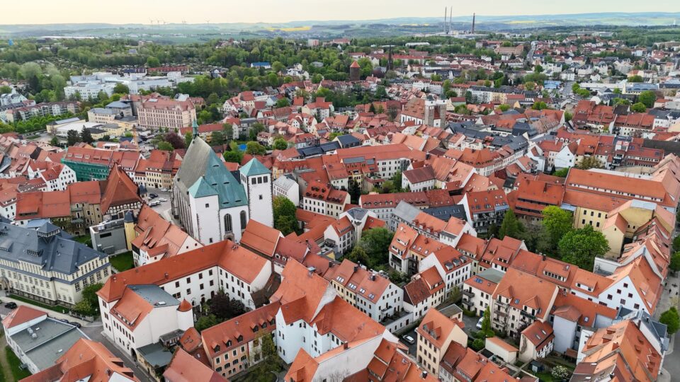 Luftaufnahme der historischen europäischen Stadt Freiberg mit roten Ziegeldächern. Auffällige Merkmale sind eine große Kirche mit zwei hohen Türmen und eine Mischung aus Wohn-, Geschäfts- und Industriegebäuden. Umgeben von Grün und fernen Hügeln unter einem klaren Himmel.