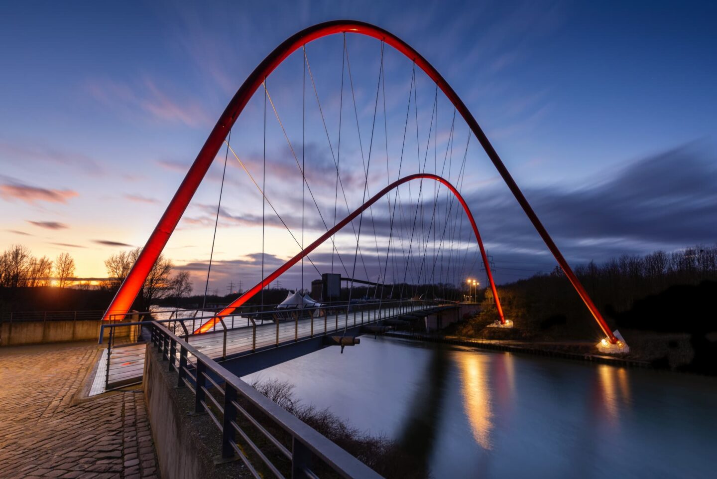 Eine moderne Brücke mit zwei großen roten Bogenpfeilern überspannt in der Abenddämmerung einen Fluss in Gelsenkirchen. Der Himmel zeigt bei Sonnenuntergang einen Farbverlauf von Blau nach Orange. Die Brücke und ihre Spiegelung auf dem Wasser erzeugen eine symmetrische, optisch beeindruckende Szene.