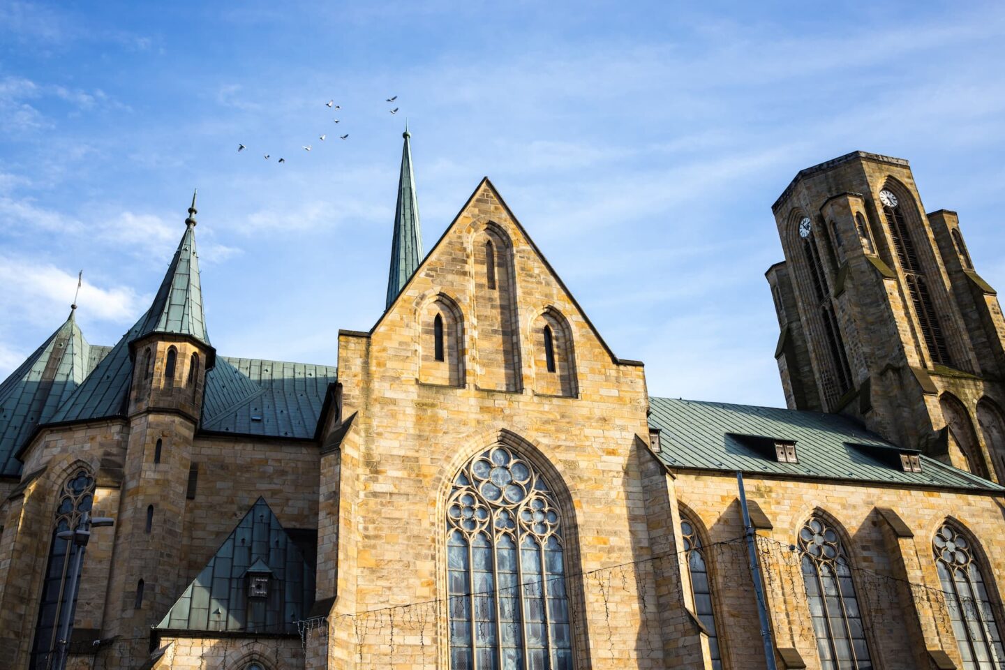 Eine historische Kathedrale mit Steinfassade und hohen, spitzen Türmen unter einem strahlend blauen Himmel schmückt das Herz von Gelsenkirchen. Das Gebäude verfügt über große Bogenfenster und komplizierte architektonische Details. Vögel fliegen über einem der Kirchtürme, während die Sonne Schatten auf das Bauwerk wirft.