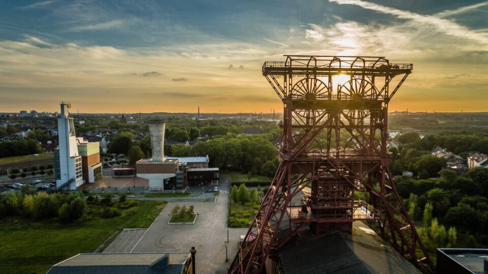 Luftaufnahme eines Industriegeländes in Gelsenkirchen mit einer großen Metallkonstruktion im Vordergrund, möglicherweise ein Fördergerüst, bei Sonnenuntergang. Gebäude und üppiges Grün umgeben das Gebiet, in der Ferne ist die Skyline der Stadt unter einem teilweise bewölkten Himmel zu sehen.