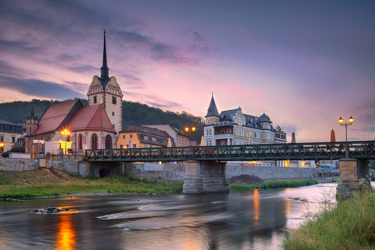 Eine ruhige Stadtlandschaft bei Sonnenuntergang mit einer Steinbrücke über einem ruhigen Fluss in Gera. Auf der einen Seite steht eine Kirche mit einem hohen Kirchturm zwischen anderen historischen Gebäuden. Der Himmel ist in Rosa- und Lilatönen gemalt und die Straßenlaternen entlang des Flussufers strahlen ein warmes Licht aus.