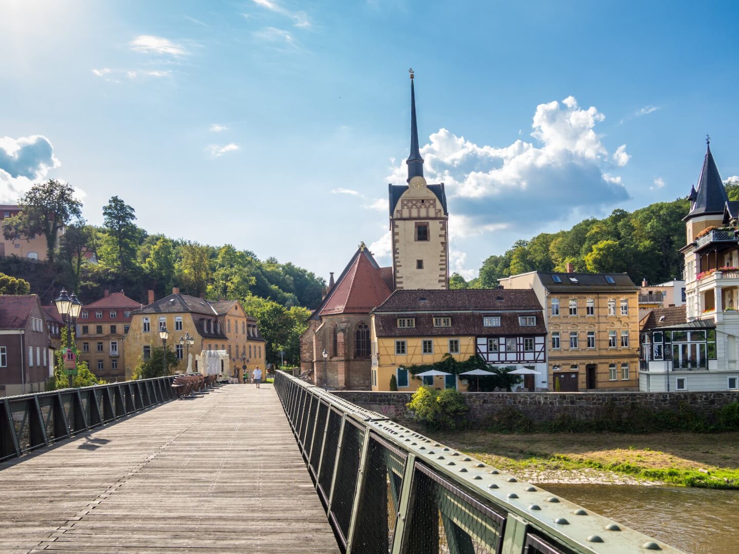 Eine malerische Szene einer kleinen europäischen Stadt, die an Gera erinnert, mit einer Holzbrücke, die zu malerischen historischen Gebäuden und einer Kirche mit einem hohen Turm führt. Ein strahlend blauer Himmel mit vereinzelten Wolken und üppigen grünen Bäumen im Hintergrund vervollständigen die idyllische Landschaft.