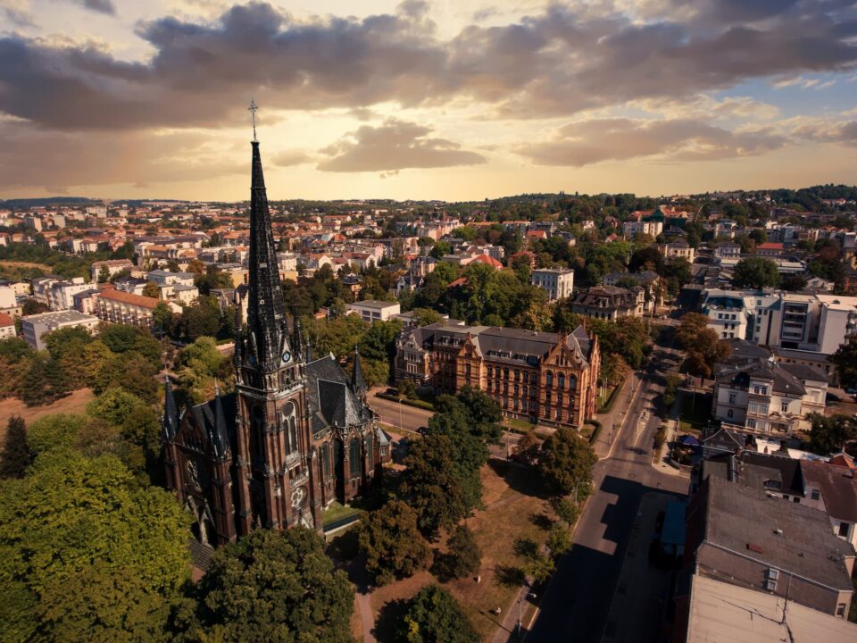 Luftaufnahme einer gotischen Kirche mit hohem Turm in Gera, umgeben von üppigen grünen Bäumen in einer Stadtlandschaft bei Sonnenuntergang. Die Skyline umfasst eine Mischung aus historischen und modernen Gebäuden unter einem teilweise bewölkten Himmel.