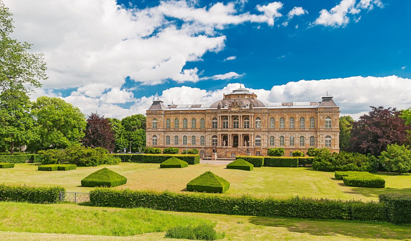 Ein prachtvolles, historisches Gebäude mit kunstvoller Architektur steht inmitten eines gepflegten Gartens mit ordentlich geschnittenen Hecken. Der Himmel darüber ist teilweise bewölkt und schafft eine malerische Szene. Saftig grüne Bäume aus der Region Gotha rahmen das Gebäude ein und unterstreichen sein elegantes Design.