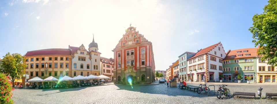 Ein Panoramablick auf einen sonnenbeschienenen Stadtplatz in Gotha zeigt farbenfrohe historische Gebäude mit Ziegeldächern. Ein zentrales, reich verziertes Gebäude sticht hervor, flankiert von Cafés mit Sitzgelegenheiten im Freien auf der linken Seite. Fahrräder und Bäume säumen den Platz und sorgen für eine lebendige Atmosphäre.