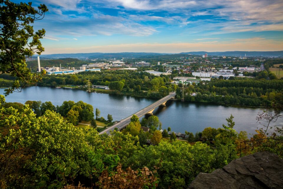 Ein malerischer Blick auf den Hagen, umgeben von üppigen grünen Bäumen und einer Brücke, die ihn überquert. Im Hintergrund eine weitläufige Stadtlandschaft mit verschiedenen Gebäuden und einer Hügellandschaft unter einem teilweise bewölkten Himmel.