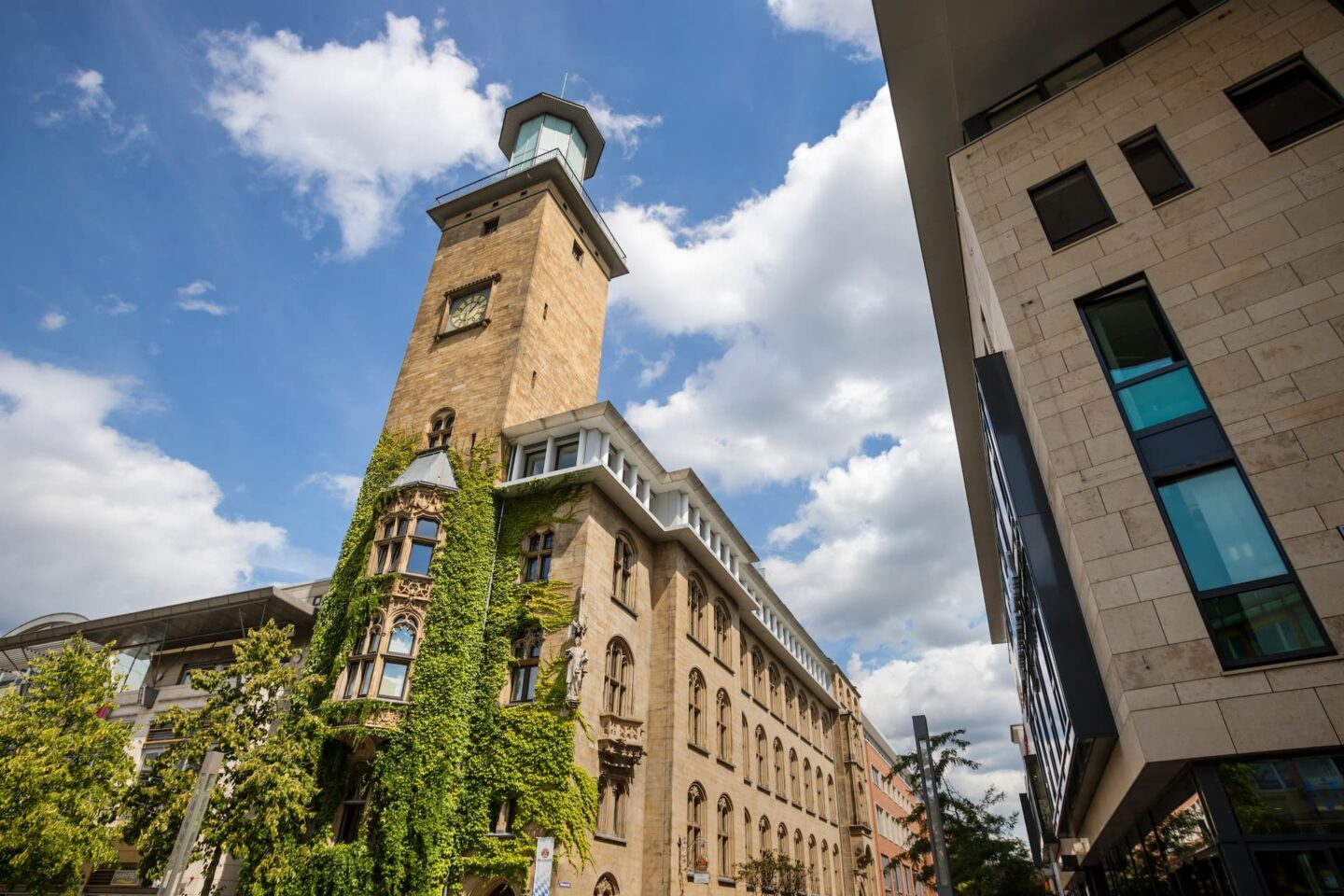 Historisches Hagen-Gebäude mit einem hohen Turm, der teilweise mit grünem Efeu bedeckt ist, vor einem strahlend blauen Himmel mit vereinzelten Wolken. Modernes Gebäude mit reflektierenden Fenstern auf der rechten Seite. Bäume und Straßenschilder säumen den Bürgersteig.