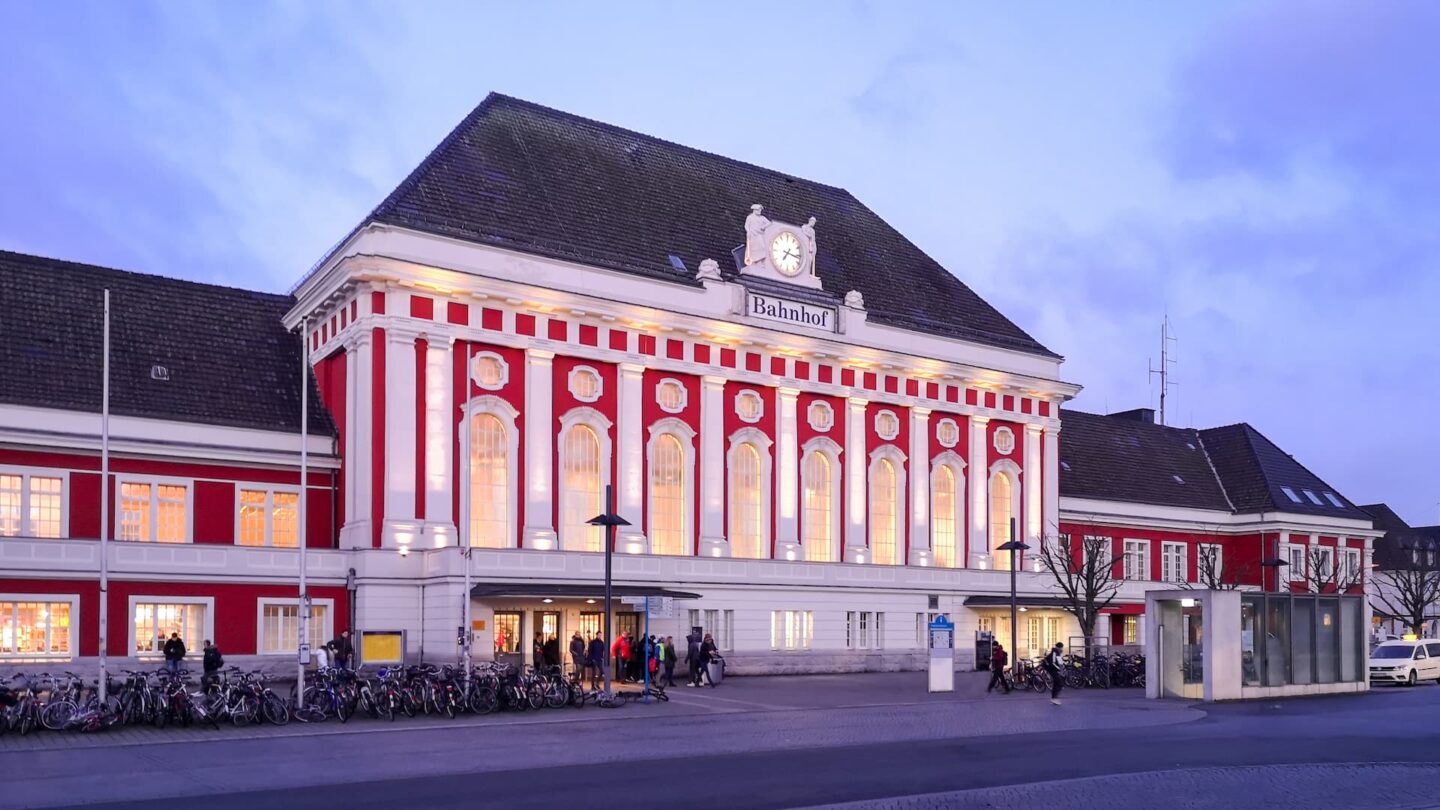 Ein großes Bahnhofsgebäude mit rot-weißer Fassade wird im Abendlicht beleuchtet. Der Mittelteil verfügt über hohe Bogenfenster und eine Uhr über dem Haupteingang. Im Vordergrund sind Fahrräder abgestellt, während sich Menschen in der Nähe des Eingangs versammeln und den Charme Hamms einfangen.