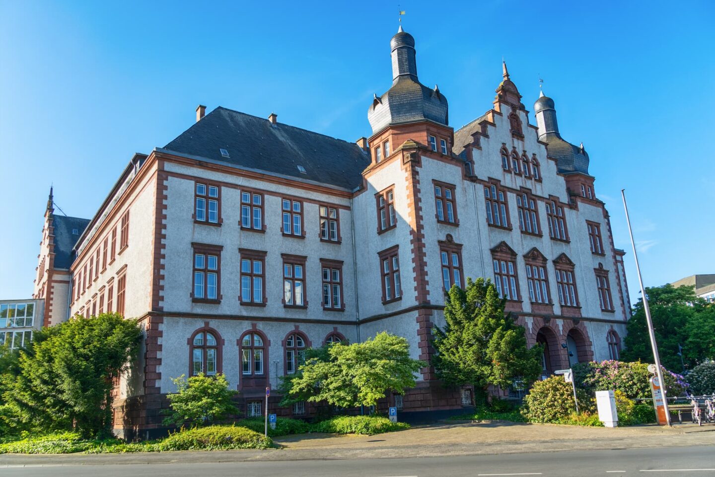 Ein großes, historisches Gebäude in Hamm mit einer Mischung aus gotischen und Renaissance-Architekturstilen, mit mehreren Bogenfenstern, zwei turmartigen Strukturen und einem steilen Dach. Das Gebäude ist von Grün umgeben und steht vor einem klaren blauen Himmel.