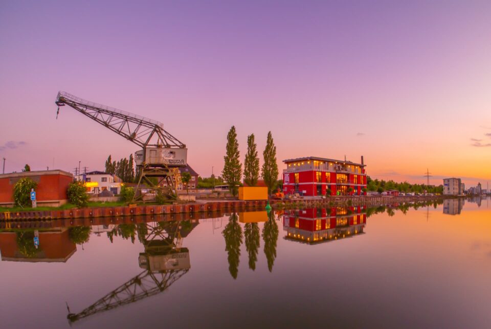 Ein ruhiges Gewässer spiegelt ein leuchtend rotes mehrstöckiges Gebäude und üppige Bäume unter einem sich verdunkelnden violett-orangefarbenen Sonnenuntergangshimmel in Hamm wider. Auf einer Seite steht ein alter Kran, der der ruhigen Szene einen industriellen Touch verleiht. Kleine Gebäude säumen ebenfalls die Uferpromenade.