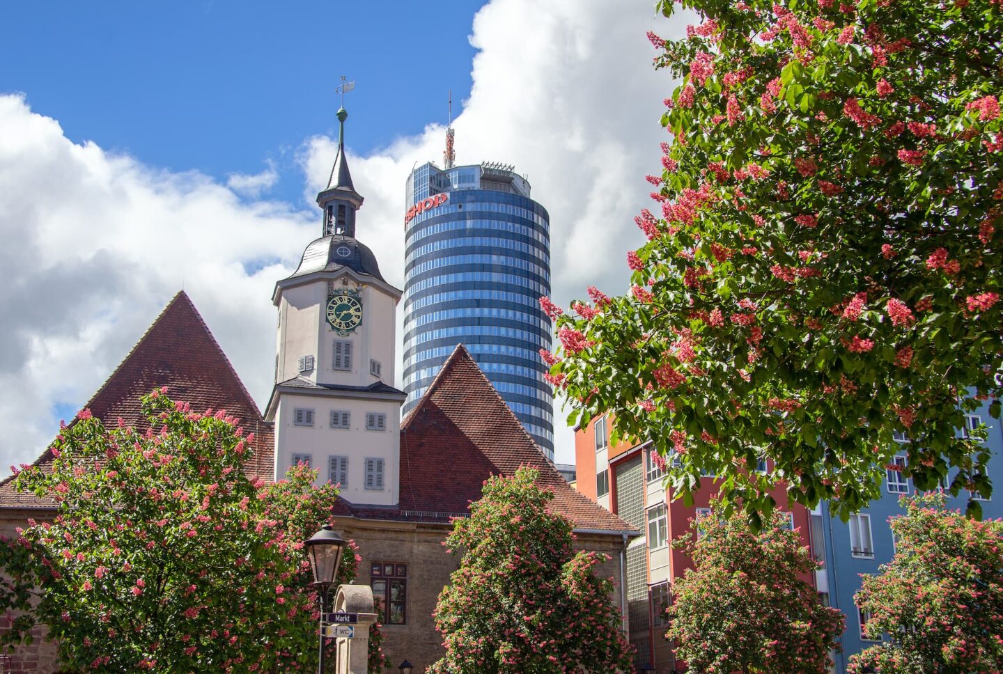 Eine malerische Szene mit einem historischen Gebäude mit einem Uhrturm und dreieckigen Dächern, umgeben von blühenden Bäumen. Im Hintergrund ragt der moderne zylindrische Wolkenkratzer von Jena mit blauen Glasfenstern vor einem teilweise bewölkten Himmel auf.