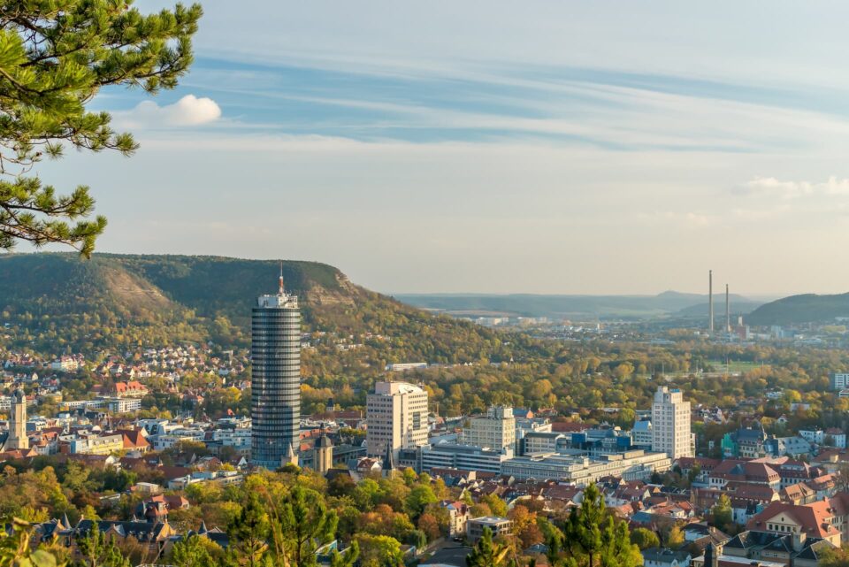 Luftaufnahme von Jena, einer Stadt umgeben von üppigem Grün und Hügeln. Hohe Gebäude, darunter ein markanter Turm, prägen die Landschaft. Der Himmel ist klar mit ein paar Wolkenstreifen. Bäume im Vordergrund rahmen die Stadtszene ein.