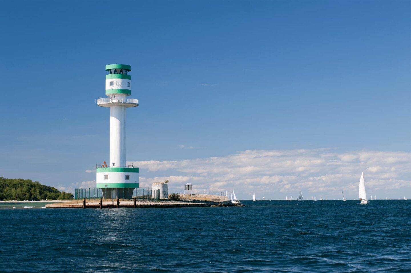 Ein grün-weißer Leuchtturm steht auf einem Betonpfeiler, umgeben vom blauen Wasser Kiels. Segelboote liegen verstreut auf dem Wasser unter einem klaren blauen Himmel. Im Hintergrund sind ein grün bewachsenes Waldgebiet und entfernte Gebäude entlang der Uferlinie zu sehen.