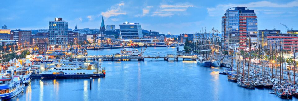 Panoramablick auf den geschäftigen Kieler Hafen in der Abenddämmerung, mit Gebäuden und Lichtern der Stadt, die sich im Wasser spiegeln. Zahlreiche Boote und Schiffe liegen am Ufer vor Anker, und im Hintergrund sind unter einem bewölkten Himmel moderne Hochhäuser zu sehen.