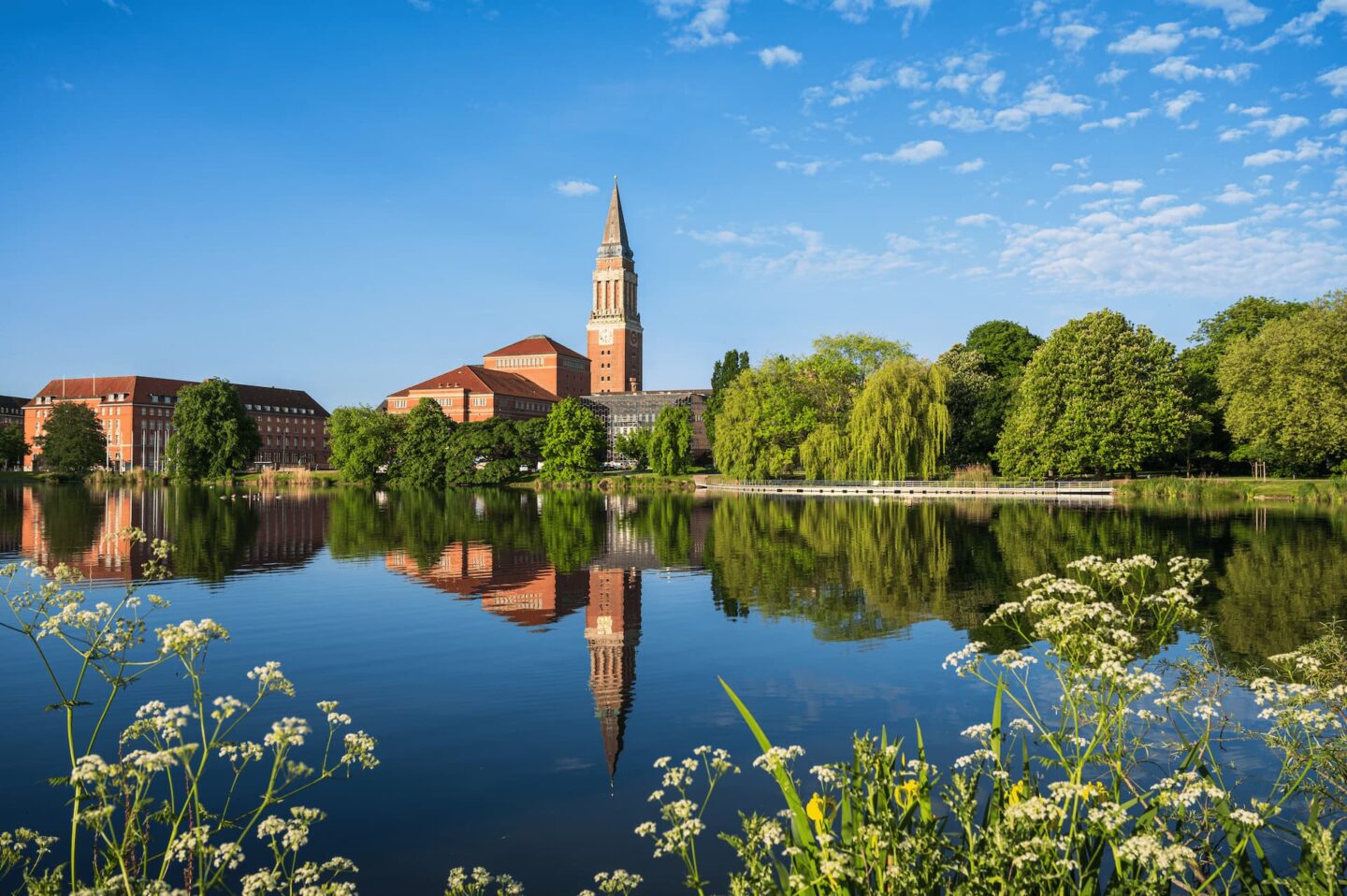 Ein großes Backsteingebäude mit einem hohen Uhrturm steht am Rande eines ruhigen Kieler Sees und spiegelt sich deutlich im Wasser. Die Szene wird von üppigen grünen Bäumen und weißen Wildblumen im Vordergrund eingerahmt. Ein strahlend blauer Himmel mit vereinzelten Wolken vervollständigt die ruhige Kulisse.