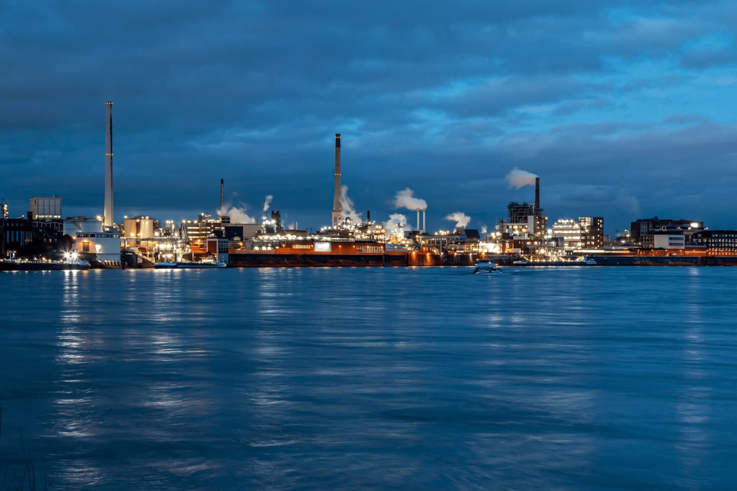 Nächtliche Industrielandschaft von Krefeld mit Fabriken und Gebäuden, die von hellen Lichtern beleuchtet werden und sich im ruhigen Wasser eines Flusses im Vordergrund spiegeln. Schornsteine stoßen weißen Rauch vor einem dunklen, bewölkten Himmel aus. Die Szene strahlt eine ruhige und doch geschäftige Atmosphäre aus.