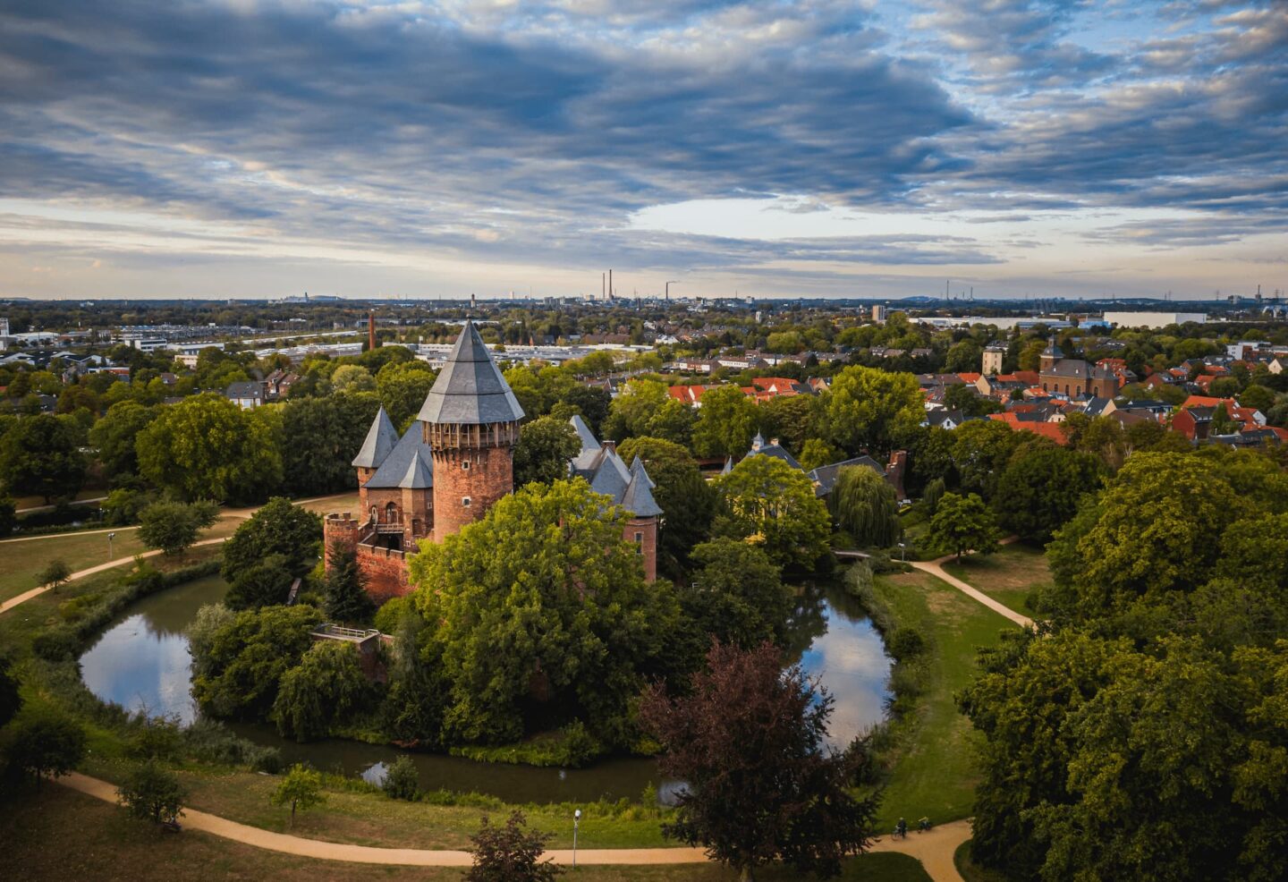 Luftaufnahme eines historischen Backsteinschlosses in Krefeld, umgeben von einem Wassergraben und üppigem Grün, mit einer malerischen Stadt und einem weiten Horizont im Hintergrund unter einem bewölkten Himmel bei Sonnenuntergang.