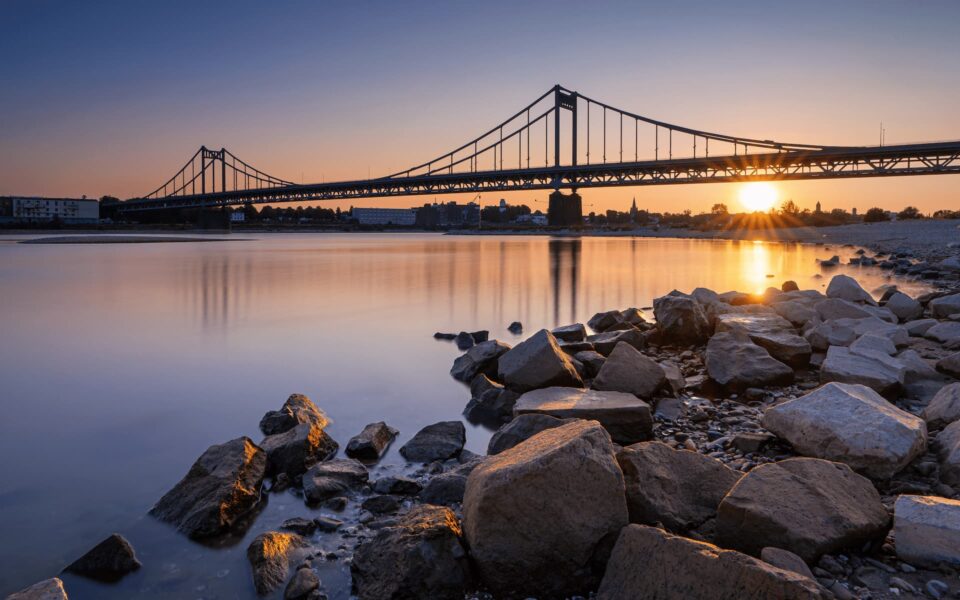 Bei Sonnenuntergang überspannt eine Hängebrücke einen ruhigen Fluss in Krefeld und wirft Spiegelbilder auf das Wasser. Der Himmel ist in warmen Orange- und Blautönen gehalten. Im Vordergrund säumen große Felsen das Ufer und verleihen der ruhigen Landschaft Struktur.