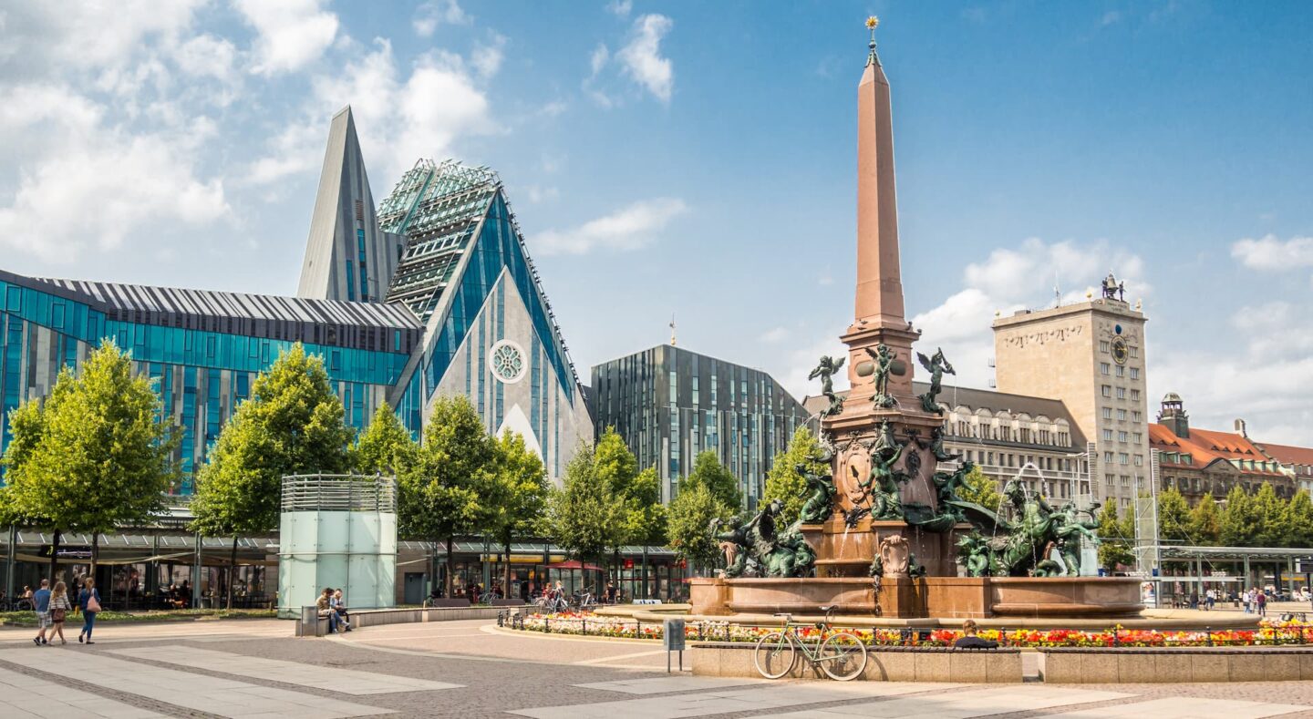 Ein lebhafter Stadtplatz in Leipzig, mit dem kunstvollen Mende-Brunnen im Vordergrund und der modernen Glasarchitektur der Universität Leipzig im Hintergrund. An einem sonnigen Tag schlendern die Menschen auf diesem bezaubernden Leipziger Platz umher und entspannen sich.