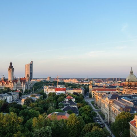 Ein Panoramablick auf Leipzig bei Sonnenuntergang zeigt eine Mischung aus modernen und historischen Gebäuden, darunter ein hohes Hochhaus und eine historische Kuppelkonstruktion. Im Vordergrund sind grüne Bäume und Wohnhäuser zu sehen, während der Himmel teilweise klar ist und sanftes Abendlicht bietet.